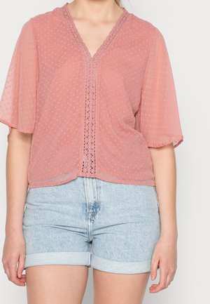 Woman wearing a sheer pink blouse with lace trim and textured dots, paired with light blue rolled-up denim shorts.
