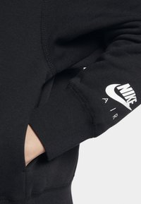 Svart sweatshirt med ribbade muddar. Har en vit Nike-logotyp och texten "AIR" på ärmen. Materialet verkar mjukt och texturerat.
