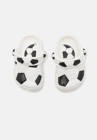 Tamancos brancos com um design de bola de futebol preto e branco, com uma sola de borracha texturizada e detalhes pretos na alça e nas laterais.