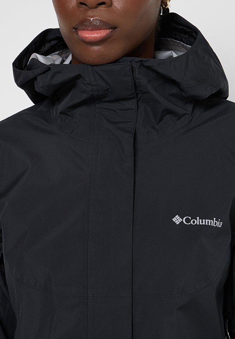 Gros plan sur une personne portant une veste à capuche noire Columbia avec un col haut et un logo visible sur la poitrine.