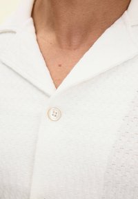 Blusa bianca in tessuto testurizzato con colletto morbido e un singolo bottone simile a una perla allo scollo, che mette in mostra un motivo sottile.