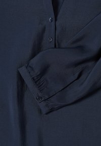 Chemise bleu marine avec des boutons, texture lisse et manches repliées. Le tissu semble léger avec un subtil éclat.