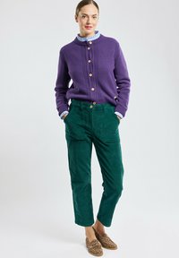 Cardigan a maglia viola con bottoni in legno, abbinato a pantaloni in corduroy verdi; presenta una camicia blu con colletto sotto e pantofole con stampa leopardata.