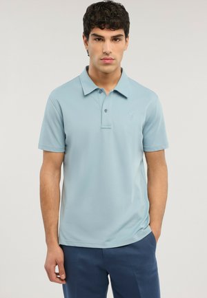 Mand iført lyseblå polo og mørkeblå bukser, stående med den ene hånd i lommen mod en ensfarvet baggrund.