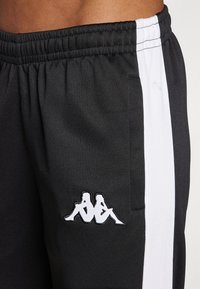 Zwarte sportbroek met een witte zijstreep. Kenmerk is een geborduurd Kappa-logo in het wit. Elastische tailleband met een zak aan de zijkant.