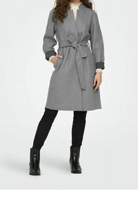Grauer Wollmantel mit langen Ärmeln, offenem Vorderteil, tailliertem Schnitt und zwei seitlichen Taschen; getragen über schwarzen Skinny-Jeans und Stiefeletten.