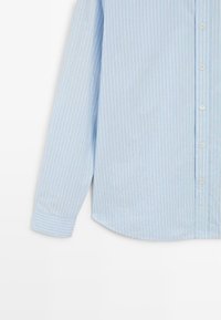 Camicia azzurro chiaro con righe verticali, apertura con bottoni e maniche lunghe. Presenta un orlo arrotondato e una finitura in tessuto testurizzato.