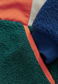 Fleece bunda s textúrami v zelených, modrých a oranžových odtieňoch. Tkanina je kožušinová s rebrovanými rukávmi a oranžovým lemovaním pozdĺž švov.