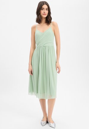 Woman with shoulder-length dark hair wearing a sleeveless mint green midi dress and silver pointed shoes, standing against a white background.