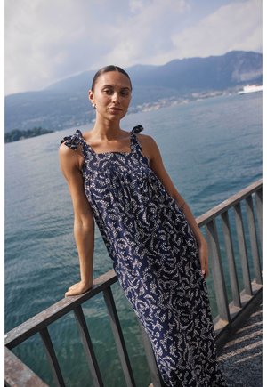 Młoda kobieta w granatowej sukience bez rękawów z białymi wzorami opiera się o metalową balustradę nad jeziorem, w tle góry w pochmurny dzień.