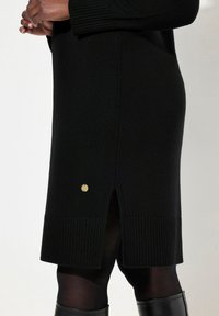 Robe en tricot noir avec un ourlet et des poignets côtelés, présente un détail doré et une fente sur le côté pour plus de mouvement et de style.