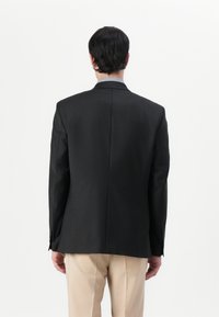 Blazer negro entallado con una textura suave, ventilación trasera sencilla y mangas largas. Presenta puños abotonados y se lleva sobre pantalones beis.