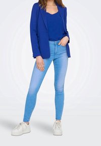 Kvinna som bär en blå kavaj och topp med ljusblå slim fit-jeans och vita sneakers med platå, stående med en hand i fickan.