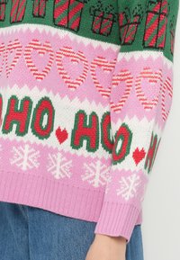 Juletrøje med grønne, pinke og hvide mønstre, der har røde og hvide hjerter, gavekasser, snefnug og "HO HO HO" tekst i rødt og grønt.