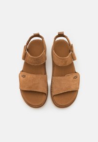 Sandales en daim marron à semelle plate, avec des brides qui se chevauchent et une fermeture réglable à la cheville. Logo brodé sur la bride.