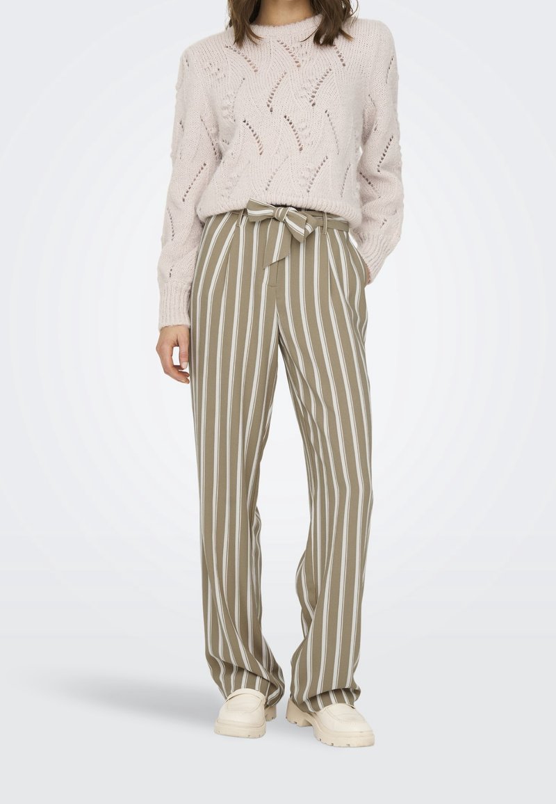 Femme portant un pull en tricot beige rentré dans un pantalon large beige et blanc à rayures verticales avec une ceinture nouée et des chaussures beiges.