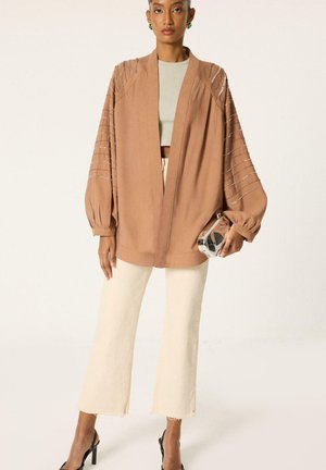 Frau trägt eine lockere braune Jacke mit Perlendetails, cremefarbene kurze Hosen, grüne Ohrringe, schwarze High Heels und hält eine silberne Clutch.
