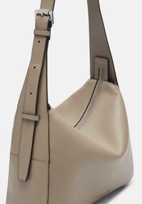Beżowa skórzana torba na ramię o zaokrąglonym kształcie, z regulowanym paskiem i detalem zamka, charakteryzująca się gładką teksturą i minimalistycznym stylem.