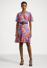 Vestido floral envolvente com mangas curtas e evasê, padrões vibrantes em vermelho, azul e laranja. Usado com um cinto trançado castanho e sandálias de plataforma.