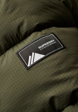 Groene pufferjack met een geruit textuur. Heeft een zwart label met de tekst "SUPERDRY" en een grafisch element in het wit.