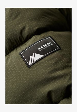 Groene pufferjack met een geruit textuur. Heeft een zwart label met de tekst "SUPERDRY" en een grafisch element in het wit.