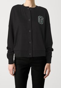 Zwarte, lichtgewicht cardigan met een ronde hals, knopen aan de voorkant en een logo op de linkerborst met het jaartal "1969." Heeft een ontspannen pasvorm en zijzakken.