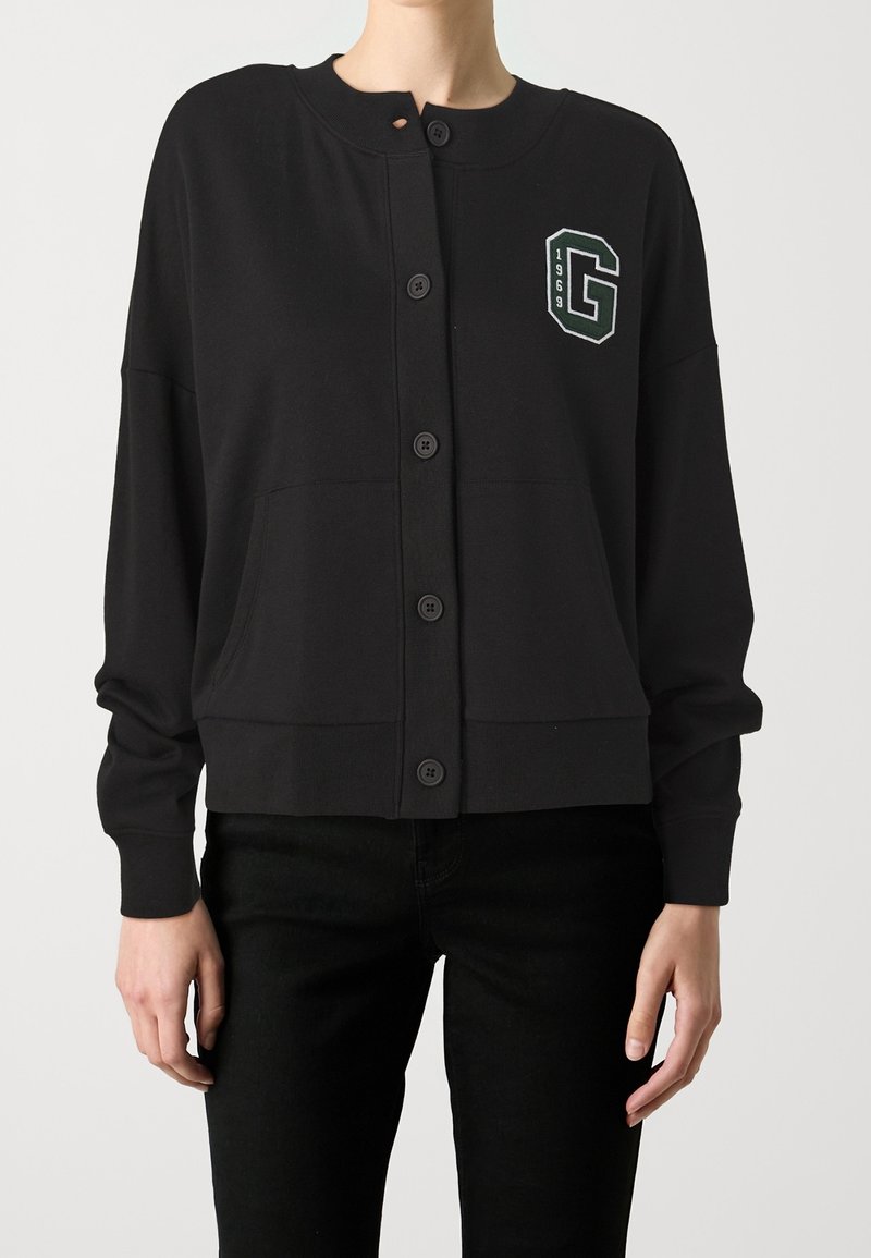 Zwarte, lichtgewicht cardigan met een ronde hals, knopen aan de voorkant en een logo op de linkerborst met het jaartal "1969." Heeft een ontspannen pasvorm en zijzakken.