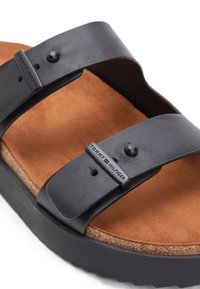 Sandale en cuir noir avec deux sangles ajustables, semelle intérieure en daim marron, semelle intermédiaire en liège et semelle noire. Logo Tommy Hilfiger sur les boucles des sangles.