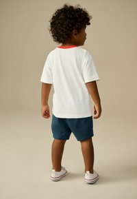 T-shirt blanc à manches courtes avec col rouge, associé à un short bleu marine et des sneakers blanches avec des accents rouges. L'enfant est montré de dos.