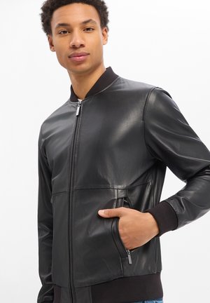 Junger Mann, der eine schwarze Lederjacke mit Reißverschluss trägt, mit gerippten Bündchen und Kragen, die Hand in der seitlichen Reißverschlusstasche vergraben, vor einem schlichten weißen Hintergrund.