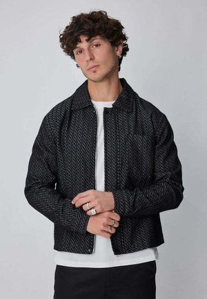 Jeune homme aux cheveux bouclés portant une veste noire texturée par-dessus une chemise blanche, debout devant un fond gris clair uni.