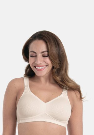 Lachende vrouw met gesloten ogen, draagt een beige draadloze beha met een textuur en bloemenborduursels op de bandjes, tegen een effen achtergrond.