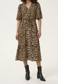 Robe portefeuille à imprimé léopard dans des tons marron clair et noir, mi-longue, avec des manches courtes, taille nouée et un tissu doux et texturé. Bottines noires.