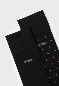 Schwarze Socken mit einer strukturierten, gerippten Oberseite, eine einfarbig und die andere mit einem Muster aus gelben Punkten und "BOSS"-Logo in Grau.