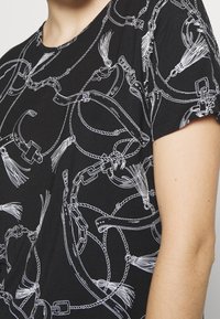 Chemise noire avec motif de chaîne blanche, ceinture, gland et corde portée par une personne montrant une partie du cou et du bras.