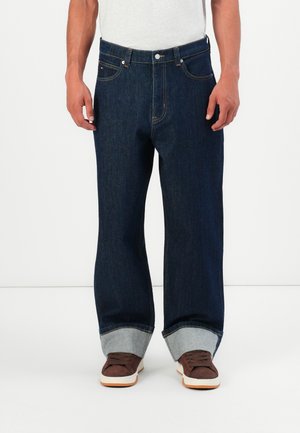 Man in een donkerblauwe wijde jeans met omgeslagen zoom en bruine sneakers, staande voor een witte achtergrond.