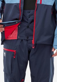 Navy blue waterproof jacket with red accents, zippered and snap closures, and inner pockets; coordinating navy pants with zippers and logo detail.