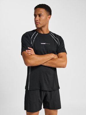 Mann steht mit verschränkten Armen, trägt ein schwarzes Sportshirt und Shorts mit weißen Paspeln vor einfarbigem Hintergrund.