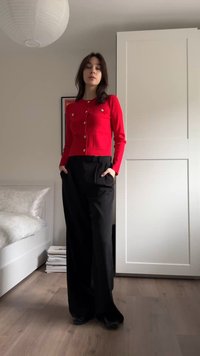 Cardigan roșu cu nasturi aurii, asortat cu pantaloni negri largi cu buzunare laterale. Podea din lemn deschis și mobilă albă în fundal.