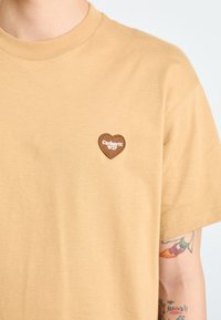 Brown short-sleeve t-shirt made of soft cotton, featuring a small embroidered heart-shaped logo in dark brown with white text on the chest.