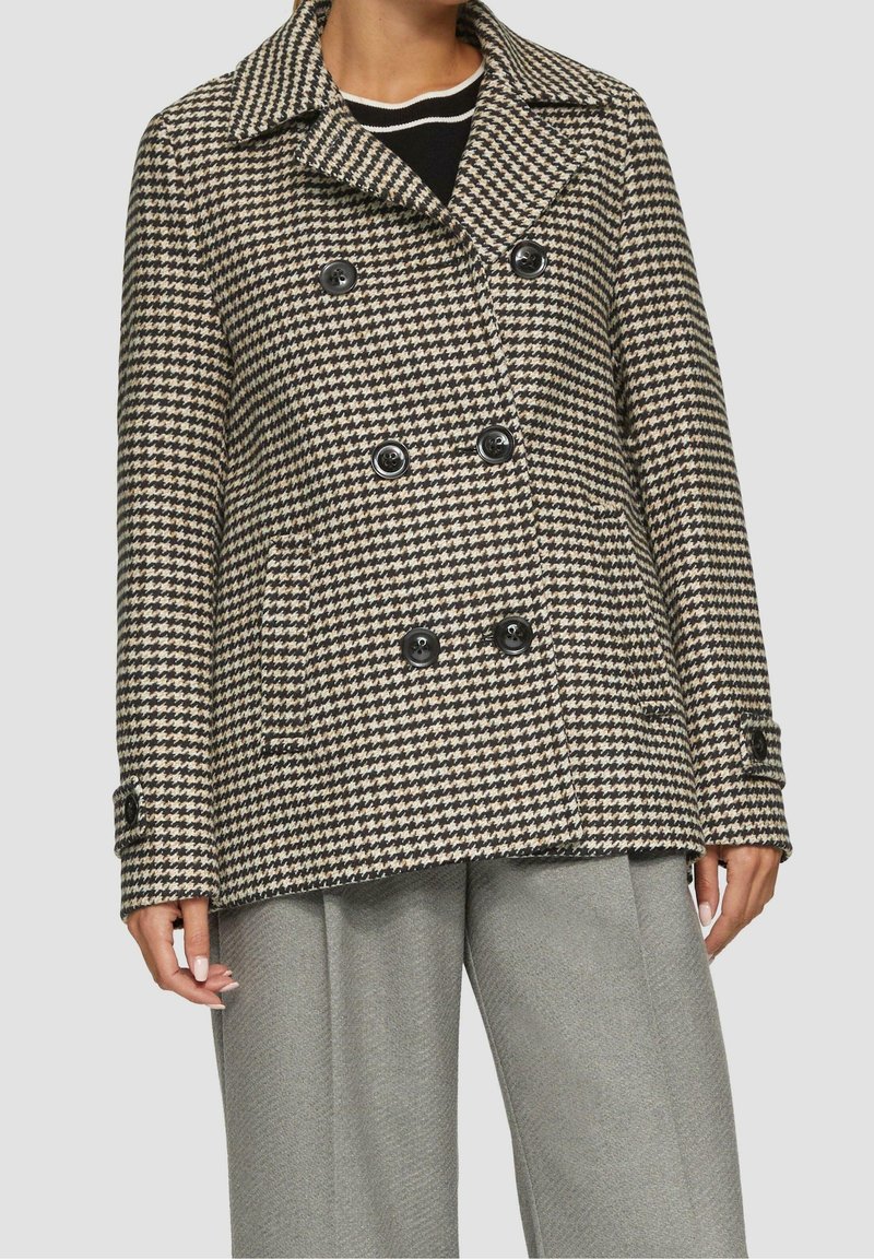 Femme portant un manteau croisé noir et blanc à motif pied-de-poule avec des boutons noirs et un pantalon gris à jambes larges.