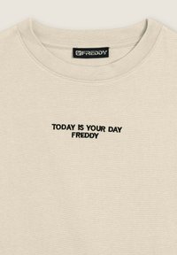 Camiseta beige de manga corta con cuello redondo. Presenta texto bordado en negro "HOY ES TU DÍA FREDDY" en el frente. Textura suave.
