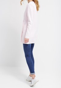 Ljusrosa lång kappa med rak design, sidofickor, i kombination med blå smala jeans och vita sneakers. Slätt, mjukt material.