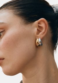 Guld- och silverhoop-örhängen med en slät, reflekterande yta. D-formad design som fästs direkt vid örsnibben.