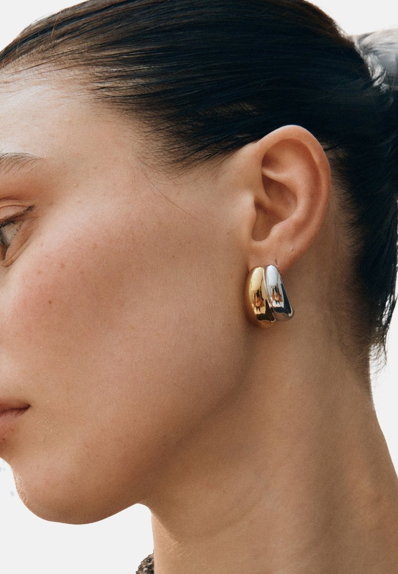 Guld- och silverhoop-örhängen med en slät, reflekterande yta. D-formad design som fästs direkt vid örsnibben.