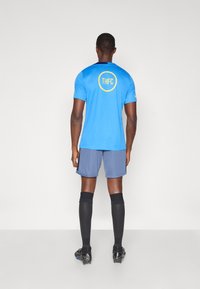 Mies, joka pitää sinistä urheilupaitaa, jossa on THFC-logo selässä, harmaita shortseja, mustia polven ylle ulottuvia sukkia ja mustia nappulakenkiä, seisoo tasaisella taustalla.