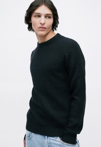 Νεαρός με μεσαίου μήκους σκούρα μαλλιά, φορώντας πουλόβερ με υφή σε σκούρο χρώμα και ανοιχτό μπλε τζιν, κοιτάζοντας πλάγια σε απλό φόντο.