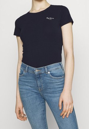 Osoba nosząca dopasowaną czarną koszulkę Pepe Jeans i niebieskie, wysokie dżinsy na jednolitym jasnym tle.