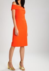 Orange Off-Shoulder Kleid mit figurbetontem Design und knielangem Saum. Getragen mit metallisch goldenen Blockabsatzsandalen. Glatte Textur.