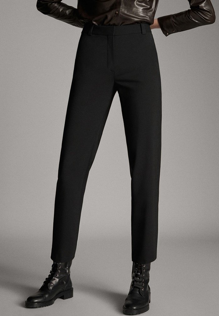 Personne portant un pantalon noir sur mesure, des bottes de combat noires à lacets et une chemise en cuir marron foncé boutonnée, les mains sur les hanches.